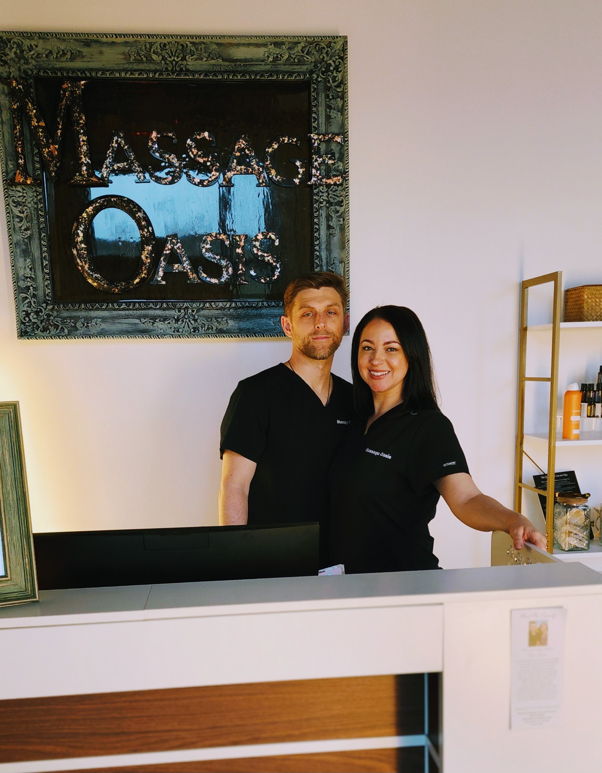 Two of our therapists in scrubs smiling behind the front desk beneath the Massage Oasis sign